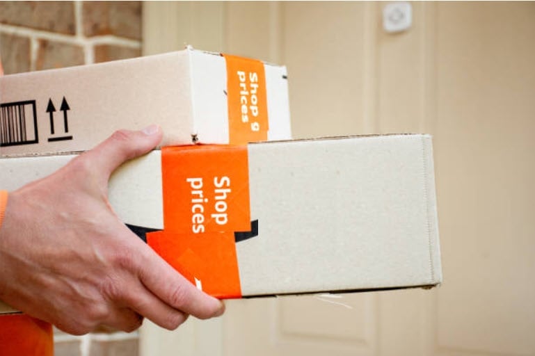 Person holding a cardboard box with orange Shop Prices shipping label against a light colored wall